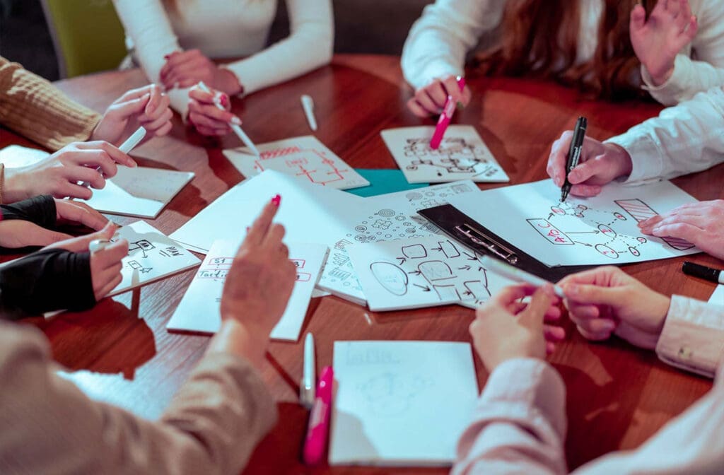Group of people brainstorming around a table, sketching marketing ideas on paper.