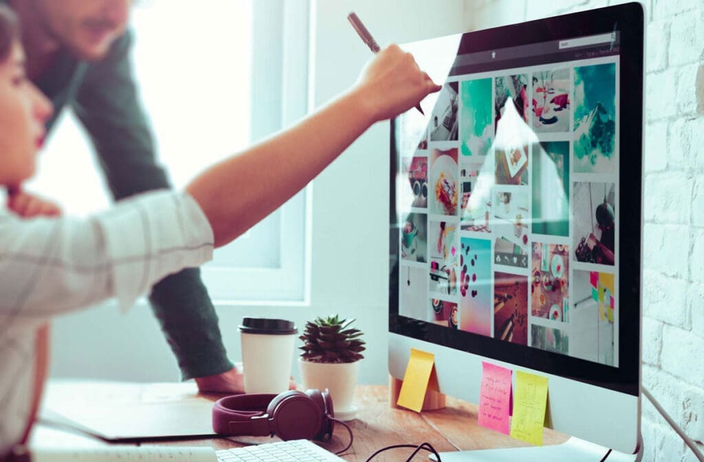 Two team members pointing at a computer screen displaying various design thumbnails.