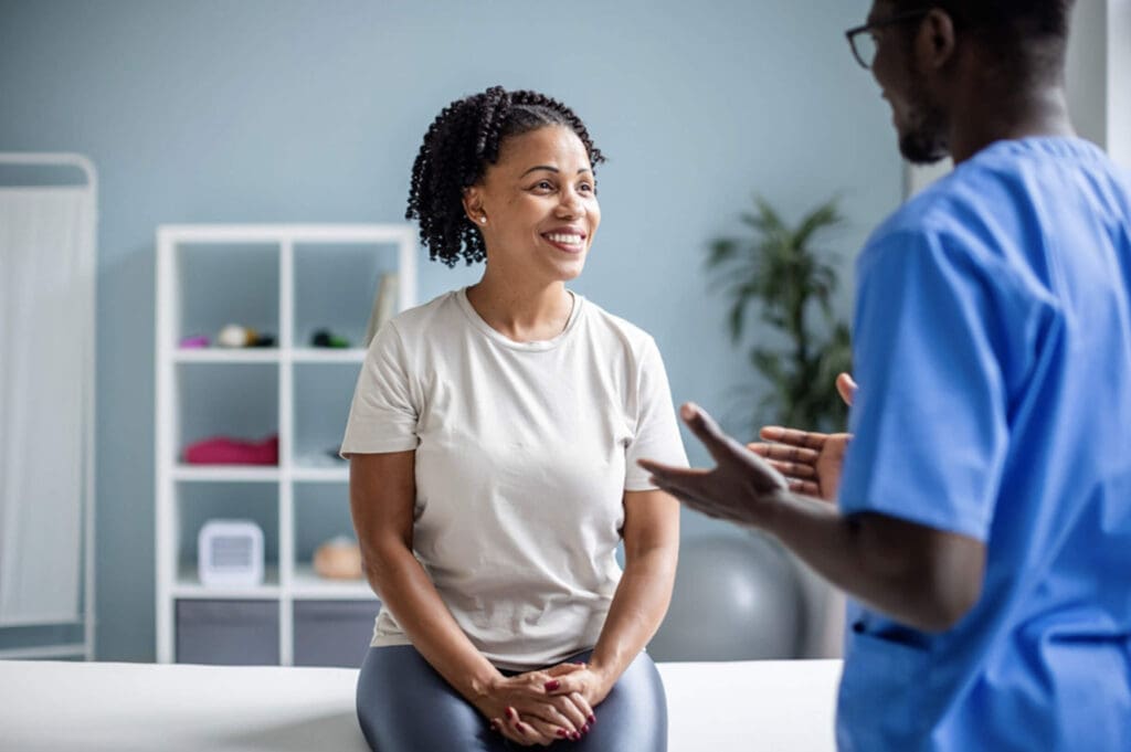 Smiling female patient sits on an exam-table while a healthcare professional in blue scrubs speaks with her inside a bright, modern exam room.